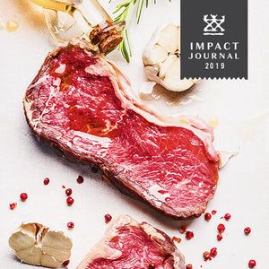 An overhead image of a raw piece of red meat drizzled with cooking oil and surrounded by peppercorns, garlic and rosemary.