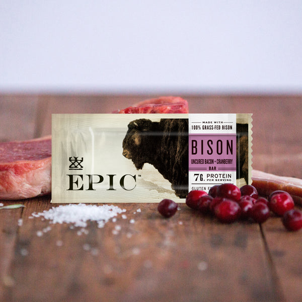 EPIC's Grass-fed Bison Bacon Cranberry bar surrounded by the ingredient included in the bar. Like salt, cranberries, bison on a rustic wood table.