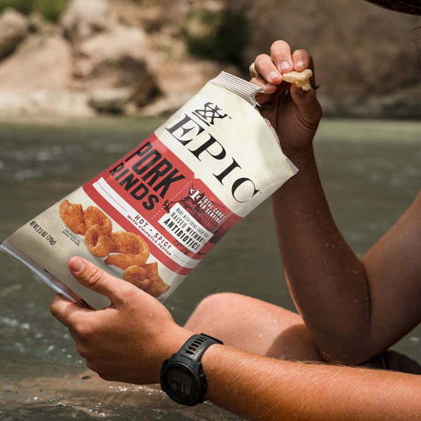 A person eating EPIC's Hot and Spicy Pork Rinds near a river.